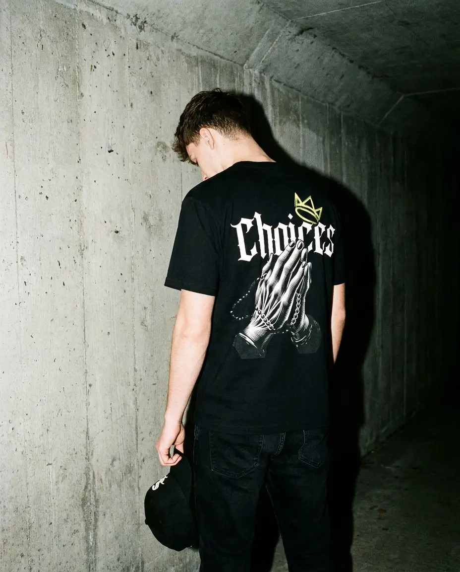 Model with head bowed against a concrete underpass wall at night in the Crowned Choices tee, praying hands back print